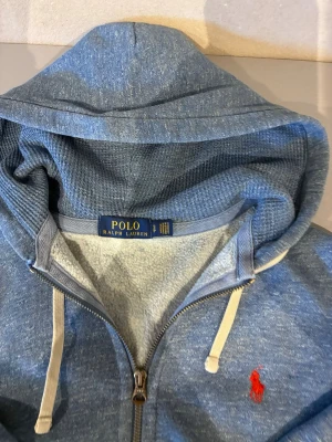 Blå hoodie från Polo Ralph Lauren - Snygg blå zip-hoodie från Polo Ralph Lauren med vit dragsko och klassisk röd logga på bröstet. Mjuk bomullsblandning, huva och fickor framtill. Perfekt för en avslappnad och stilren look.