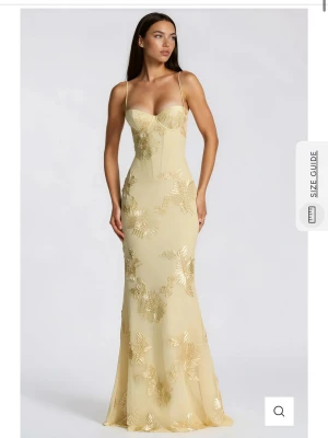 Ljus långklänning med blommigt mönster - Elegant långklänning i ljus gulfärg med tunna axelband och öppen rygg med snörning. Klänningen har blommigt mönster i guldtoner och figurnära passform. Perfekt för speciella tillfällen och sommarens fester.💛OBS:klänningen är liten i storlek