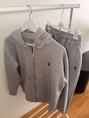  Polo Ralph Lauren - Snyggtw grått set från Polo Ralph Lauren med zip-hoodie och matchande byxor. Båda plaggen har den klassiska broderade loggan på bröstet och benet. Tillverkade i mjuk bomullsblandning för maximal komfort och avslappnad stil.