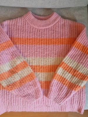 Randig stickad tröja Gina Tricot - Mysig stickad tröja från Gina Tricot i fluffigt material. Tröjan har breda ränder i rosa, orange och vitt, ribbad halsringning och muddar. Perfekt för dig som gillar färgglada och trendiga plagg med oversized känsla.
