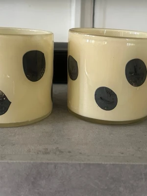 Prickiga ljuslyktor från Jakobsdals - Två ljuslyktor i glas från Jakobsdals med beige bottenfärg och stora svarta prickar runtom. Perfekta som dekoration på bordet eller i fönstret. Snygg och lekfull design som sticker ut och passar i de flesta hem.