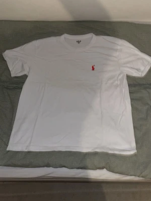 Vit t-shirt från Polo Ralph Lauren - Klassisk vit t-shirt från Polo Ralph Lauren med röd broderad logga på bröstet. T-shirten har rund halsringning och korta ärmar. Perfekt basplagg med stilren design.