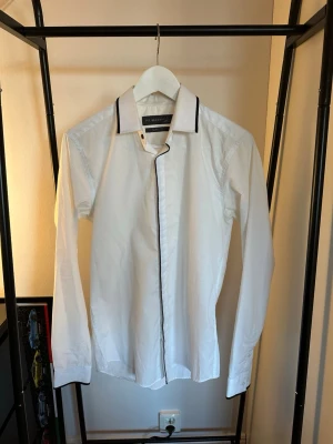 Vit skjorta med svarta detaljer Primark - Snygg vit skjorta från Primark med svarta kantdetaljer längs krage och knappslå. Klassisk passform med lång ärm och stilren design. Perfekt för dig som gillar minimalistisk stil.