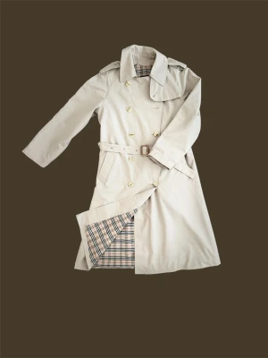 Beige trenchcoat från Burberry - Klassisk beige trenchcoat från Burberry med bälte i midjan och dubbelknäppning. Jackan har axelklaffar, stora slag och det ikoniska rutiga fodret. Perfekt för dig som vill ha en tidlös och stilren look. Materialet är bomullsmix.