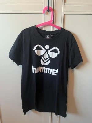 Svart Hummel t-shirt med logga - Snygg svart t-shirt från Hummel med stor vit logga framtill. Klassisk rund halsringning och korta ärmar. Tillverkad i mjuk bomull som känns skön mot huden. Perfekt för en sportig och avslappnad stil.