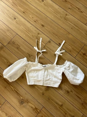 Vit croppad topp med puffärmar - Söt vit croppad topp med puffiga ärmar och knytbara axelband. Toppen har en off shoulder-design och är perfekt för en trendig och somrig look. 