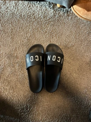 Dsquared2 ICON - Säljer ett par svarta slides från Dsquared2 med bred rem där det står 'ICON' i vit text. Sandalerna har en klassisk slip-on design och är tillverkade i syntetmaterial med räfflad sula för extra grepp. Perfekta för chill dagar eller till stranden.