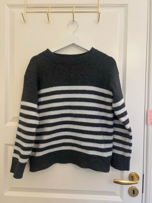 Mörkgrå stickad tröja med ränder - Mysig stickad tröja i mörkgrått med vita horisontella ränder över bröstet och ärmarna. Tröjan har rund halsringning, långa ärmar och ribbade muddar vid ärmslut och nederkant. Perfekt för kyliga dagar och enkel att matcha.