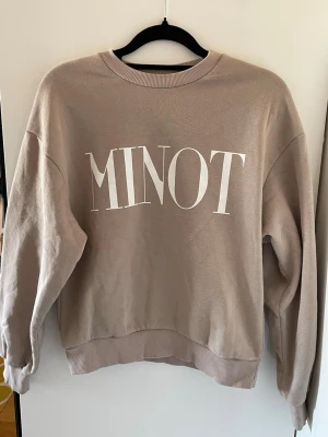 Beige sweatshirt MINOT tryck - Säljer en beige sweatshirt från Ginatricot med stort vitt tryck på bröstet. Tröjan har rund halsringning, ribbade muddar vid ärmslut och nederkant samt en avslappnad passform. Perfekt för en chill och stilren look.