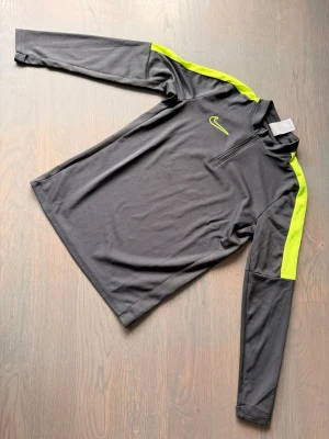 Nike långärmad träningströja grå/gul - Nike långärmad träningströja i mörkgrått med neongula detaljer på axlar och ärmar. Tröjan har halv dragkedja framtill och är tillverkad i ett lätt, funktionsmaterial som andas. Perfekt för träning eller löpning.