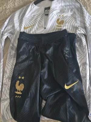 Frankrike Nike träningsset - Säljer ett träningsset från Nike för Frankrikes landslag. Setet består av en vit långärmad zip-tröja med gråa detaljer och guldiga loggor samt svarta träningsbyxor med guldiga detaljer och elastisk midja. Både tröja och byxor har Frankrikes emblem och Nike swoosh.