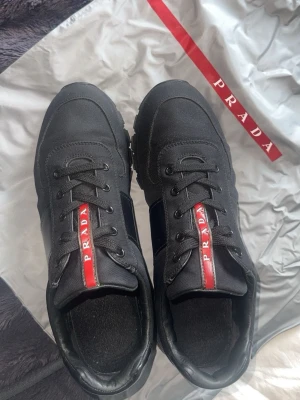 Prada sneakers med röd detalj - Snygga svarta sneakers från Prada med klassisk låg siluett.  de är storlek 42 kommer med dustbag den är ny annvödn max 1 gång pris kan diskuteras och för mer bilder bara skriva och fraktar genom plick och tar byten beror på vad det är 