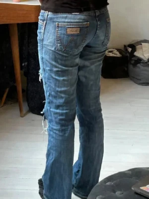 Blå slitna jeans från Wrangler - Säljer ett par blå jeans från Wrangler med raka ben och slitningar vid knäna. Jeansen har klassiska fem fickor, låg midja och råa kanter vid hålen. Perfekta för en avslappnad och trendig look.