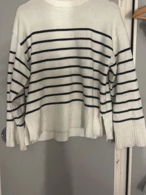 Vit stickad tröja med svarta ränder - Säljer en vit stickad tröja med svarta horisontella ränder. Tröjan har en avslappnad passform, ribbstickade muddar och nederkant samt rund halsringning. Perfekt för dig som gillar klassisk och enkel stil.