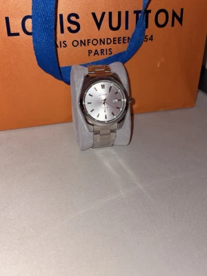 Silverfärgad klocka  - Stilren klocka  med silverfärgad länkarmband och rund urtavla. Urtavlan är vit med tydliga markeringar och datumvisning. Klassisk design som passar till många olika stilar. Metallarmbandet ger en clean och modern känsla.