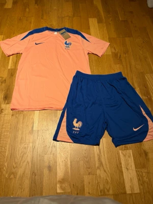 Frankrike Nike fotbollskit rosa/blå - Säljer ett Frankrike fotbollskit från Nike med en ljusrosa t-shirt och blå detaljer samt blå shorts med rosa inslag. Tröjan har FFF-logga och Nike swoosh på bröstet, shortsen har resår i midjan och samma loggor. Perfekt för träning eller match. På bilden ser det ut som orange men det är rosa egentligen.
