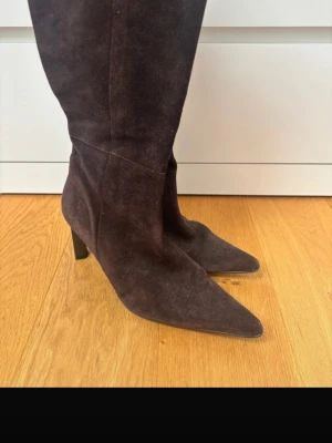 Bruna mockastövlar med klack - Snygga bruna stövlar i mocka med spetsig tå och smal klack. Stövlarna går upp till knäet och har en elegant, tidlös design som passar perfekt till höst- och vinteroutfits. Materialet är mjukt och ger en lyxig känsla.