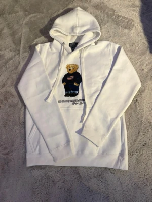 Vit hoodie med Polo Bear från Ralph Lauren - Vit hoodie från Ralph Lauren med den ikoniska Polo Bear-trycket på bröstet. Björnen har på sig en mörkblå tröja med amerikanska flaggan och jeans. Hoodien har huva med dragsko och känguruficka framtill. Perfekt för en avslappnad och trendig stil. Nu utan prislapp.
