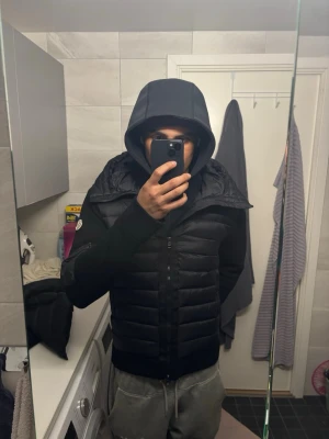 Svart pufferjacka från Moncler - Säljer en svart pufferjacka från Moncler med huva och dragkedja framtill. Jackan har stickade ärmar och detaljer samt en ficka med dragkedja på ena ärmen. Klassisk Moncler-logga på ärmen. Perfekt för kalla dagar och riktigt snygg streetstil.