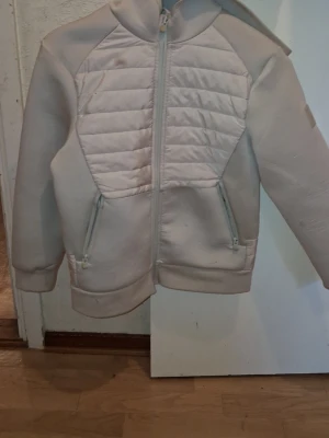 Beige quiltad jacka med huva - Snygg beige jacka med quiltade partier framtill och på huvan. Jackan har dragkedja hela vägen, två fickor med dragkedja och långärmad design. Perfekt för kyligare dagar och enkel att matcha med olika outfits.