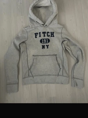 Grå hoodie från Abercrombie & Fitch med tryck - Säljer en grå hoodie från Fitch med mörkblå text 'FITCH 191 NY' på bröstet. Hoodien har en stor känguruficka framtill, dragsko i huvan och synliga sömmar för en sportig look. Perfekt för chill dagar eller när du vill ha en avslappnad stil.