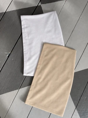 Två tubtoppar i vitt och beige - Två stilrena tubtoppar i vitt och beige. De är sydda i mjukt, stretchigt tyg och har en enkel, rak design utan axelband. Perfekta för lager på lager eller som basplagg till olika outfits. Materialet känns bekvämt och följsamt mot huden.