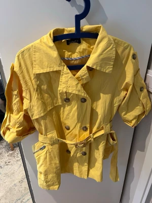 Gul trenchcoat  - Gul trenchcoat med korta ärmar, dubbelknäppning och bälte i midjan. Jackan har dekorativa knappar på axlar och ärmar samt praktiska fickor med dragkedja.  Priset går att förhandla!