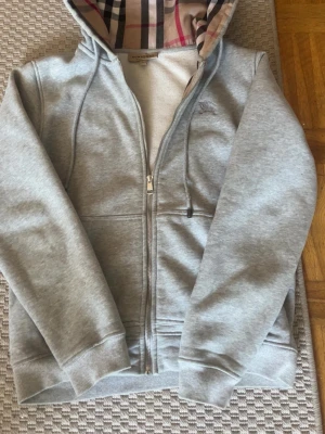 Grå zip hoodie från Burberry - Snygg grå zip hoodie från Burberry med klassiskt rutigt foder i huvan. Hoodien har dragsko i huvan, två fickor fram och ribbade muddar. Tillverkad i mjukt bomullsmaterial som känns skönt mot huden. Perfekt för en avslappnad och stilren look.