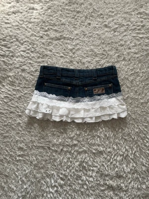 Jeanskjol med volang och spets - En Lågmidjad jeanskjol med vit volang och spetsdetaljer omsydd från ett par jeans. 🌷  Midjemått tvärsöver- 40cm Längd-27cm