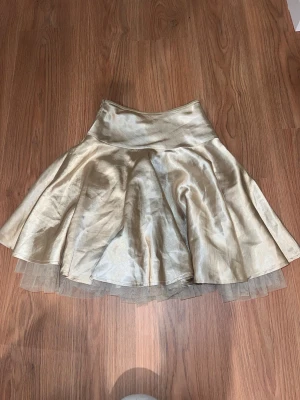 Guldig kort klänning med tyll - Snygg kort klänning i glansigt guldigt tyg med tyll underkjol för extra volym. Klänningen har en markerad midja och dragkedja i sidan för enkel påklädning. Perfekt för dig som vill sticka ut med en glittrig look.