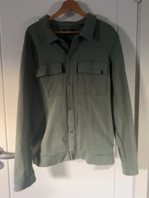 Grön overshirt med fickor - Stilren overshirt i ljusgrönt med två stora bröstfickor och knappar framtill. Skön, rak passform och klassisk krage. Perfekt att bära som lager över t-shirt. Materialet känns mjukt och lätt, troligen bomull eller bomullsblandning.