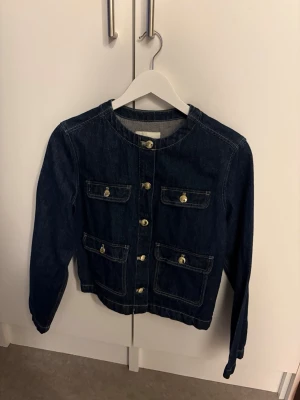 Mörkblå jeansjacka med guldknappar - Snygg mörkblå jeansjacka med rund halsringning och fyra framfickor. Jackan har dekorativa guldknappar framtill och på fickorna, samt kontrastsömmar som ger en cool look. Perfekt att slänga över en t-shirt för en avslappnad stil.