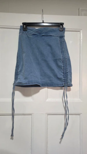 Blå kort jeanskjol med snörning - Ny & aldrig använd. Säljer en ljusblå kort jeanskjol med snörning längs sidorna och dragkedja bak. Kjolen har en tight passform och är gjord i mjukt denimtyg. Perfekt för dig som gillar en trendig och avslappnad stil.