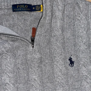 Grå kabelstickad tröja Polo Ralph Lauren - Snygg grå kabelstickad tröja från Polo Ralph Lauren med halv dragkedja och hög krage. Tröjan har det klassiska mörkblå logobroderiet på bröstet och ribbade muddar vid ärmslut och nederkant. Perfekt för lager på lager och riktigt stilren.