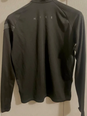 Koszulka treningowa Nike/ Träningströja Nike - Svart långärmad träningströja från Nike med halv dragkedja framtill och diskret logga på ryggen. Tillverkad i ett stretchigt funktionsmaterial som andas, perfekt för löpning eller gym. Tröjan har meshdetaljer på axlarna för extra ventilation. Denna tröja motsvarar storlek S/M. 