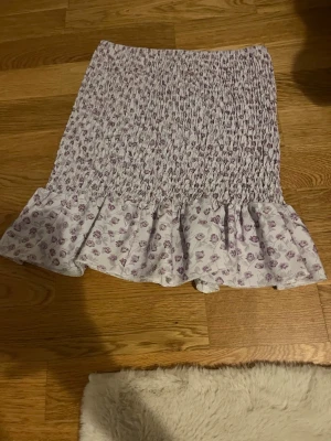 Ljuslila blommig volangtröja - Supersöt kort kjol i ljuslila med små blommor och volangkant. Kjolen har smockad midja för skön passform och är perfekt för vår och sommar. Passar till både sneakers och sandaler.