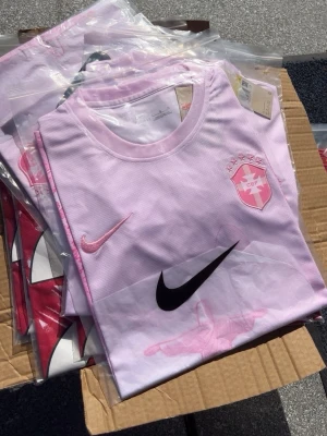 Ljusrosa Nike fotbollströja - Säljer en ljusrosa fotbollströja från Nike med rund hals och tryckt klubbmärke på bröstet. Tröjan har både rosa och svart Nike-logga samt detaljer i vitt. Perfekt för träning eller match.