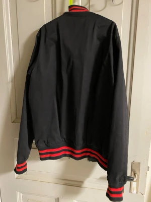 Svart bomberjacka med röda detaljer - Snygg svart bomberjacka från Dickies med dragkedja framtill. Jackan har ribbade muddar och krage med röda och svarta ränder, samt en klassisk collegejacke-look. Tillverkad i slitstarkt material, perfekt för en streetwear-stil.