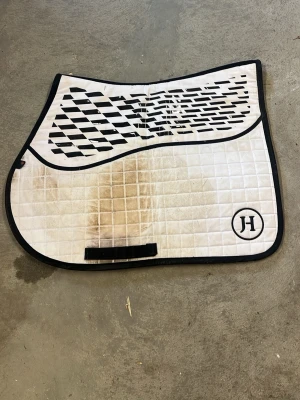 Vitt schabrak med svart kant - Säljer ett vitt schabrak med svart kant och svart/vitt grafiskt mönster upptill. Schabraket har en rundad form och är quiltat med rutmönster. Märkt med JH-logga i ena hörnet. Perfekt till hästutrustning. Behöver tvättas