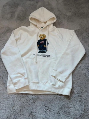 Vit hoodie Polo Bear Ralph Lauren - Vit hoodie från Ralph Lauren med den ikoniska Polo Bear-trycket på bröstet. Hoodien har snörning i huvan och en klassisk känguruficka framtill. Perfekt för dig som gillar streetwear och vill ha en stilren look. Ny utan prislapp