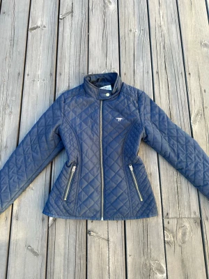 Marinblå quiltad jacka från 27 - Snygg marinblå quiltad jacka från 27 med guldfärgad dragkedja och två fickor med dragkedja. Jackan har ståkrage med tryckknapp och ett snyggt rutmönster. Den är i stolek 164 men passar också som en XS. Jackan har två dragkedjefivkor och kan någon gång kan dragkedjorna haka sig så det är smart att dra försiktigt.