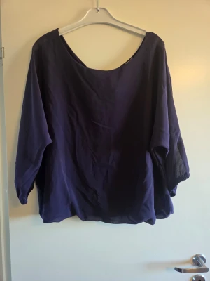 Mörklila blus med knyt i ryggen - Säljer en mörklila blus med lös passform och trekvartsärmar. Blusen har en bred urringning och en snygg knytning i ryggen som detalj. Tillverkad i ett tunt, mjukt tyg som ger ett luftigt intryck. Perfekt för dig som gillar en avslappnad men trendig stil.