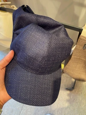 Mörkblå keps med fiskbensmönster - Snygg mörkblå keps med diskret fiskbensmönster och lätt glansig finish. Klassisk böjd skärm och sexpanelers design. Perfekt för dig som vill ha en stilren och trendig accessoar till vardagslooken.