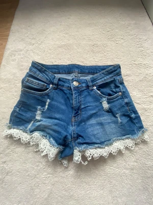 Blå jeansshorts med spetskant - Säljer ett par blå jeansshorts med snygga slitningar och vit spetskant längst ner. Shortsen har fem fickor, bälteshällor och knappstängning. Perfekta för sommaren och ger en cool, feminin vibe.
