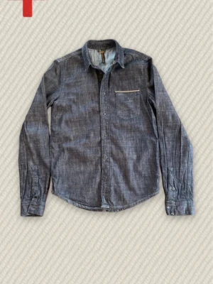 Nudie selvedge skjorta - Snygg mörkblå selvedge jeansskjorta med klassisk krage och knappstängning framtill. Skjortan har en bröstficka med vit detaljsöm och långa ärmar med manschetter. Perfekt till ett avslappnat streetwear- eller casual look.