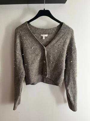 Grå kofta med pärlor från H&M - Snygg grå kofta från H&M med dekorativa pärlor över hela framsidan och ärmarna. Koftan har v-ringning, tre knappar framtill och är lite croppad i modellen. Perfekt för dig som vill ha en trendig och mysig look. En knapp sitter lösare men är enkelt att fixa, finns en extra knapp som sitter på lappen.