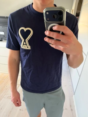 Ami Paris t-shirt  - Säljer en marinblå t-shirt från Ami Paris med en stor hjärtdetalj och bokstaven A på bröstet. T-shirten har rund hals, korta ärmar och är gjord i mjuk bomull. Perfekt för en avslappnad och trendig look.