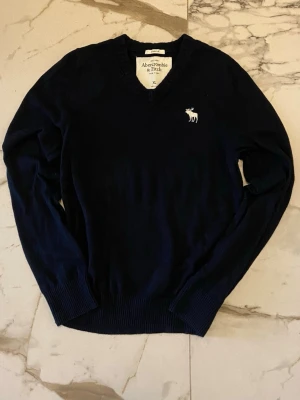 Mörkblå stickad tröja Abercrombie V Neck-M - Mörkblå stickad tröja från Abercrombie & Fitch med V-ringning och ribbade muddar. Tröjan har en vit broderad älg-logga på bröstet och är tillverkad i mjukt material. Perfekt för lager på lager och enkel att matcha. Tröjan sägs vara i storlek XL Men passar som en M!