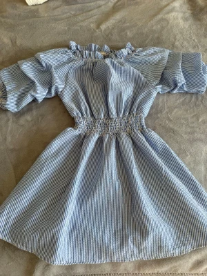 Blåvit randig kortklänning - Söt blåvit randig kortklänning med puffärmar och resår i midjan. Klänningen har volangdetaljer vid halsen och är tillverkad i ett lätt bomullstyg. Perfekt för varma dagar och ger en fräsch sommarkänsla.