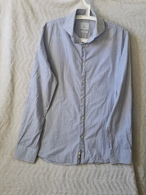 Vit och blå mönstrad skjorta från Bläck - Snygg vit skjorta från Bläck med småblått mönster över hela tyget. Skjortan har klassisk krage, knappar framtill och långa ärmar. Perfekt för dig som gillar stilrena detaljer och en modern passform.